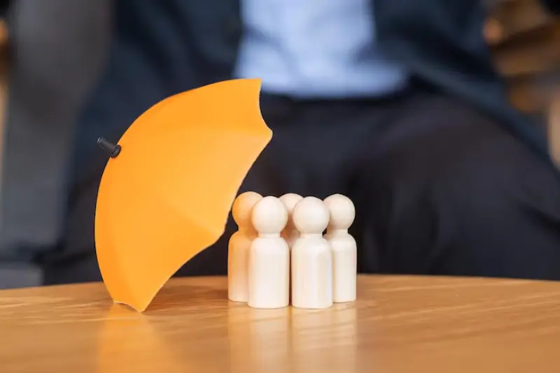 A small yellow umbrella shelters three wooden figurines, symbolizing protection and safety in a business environment.